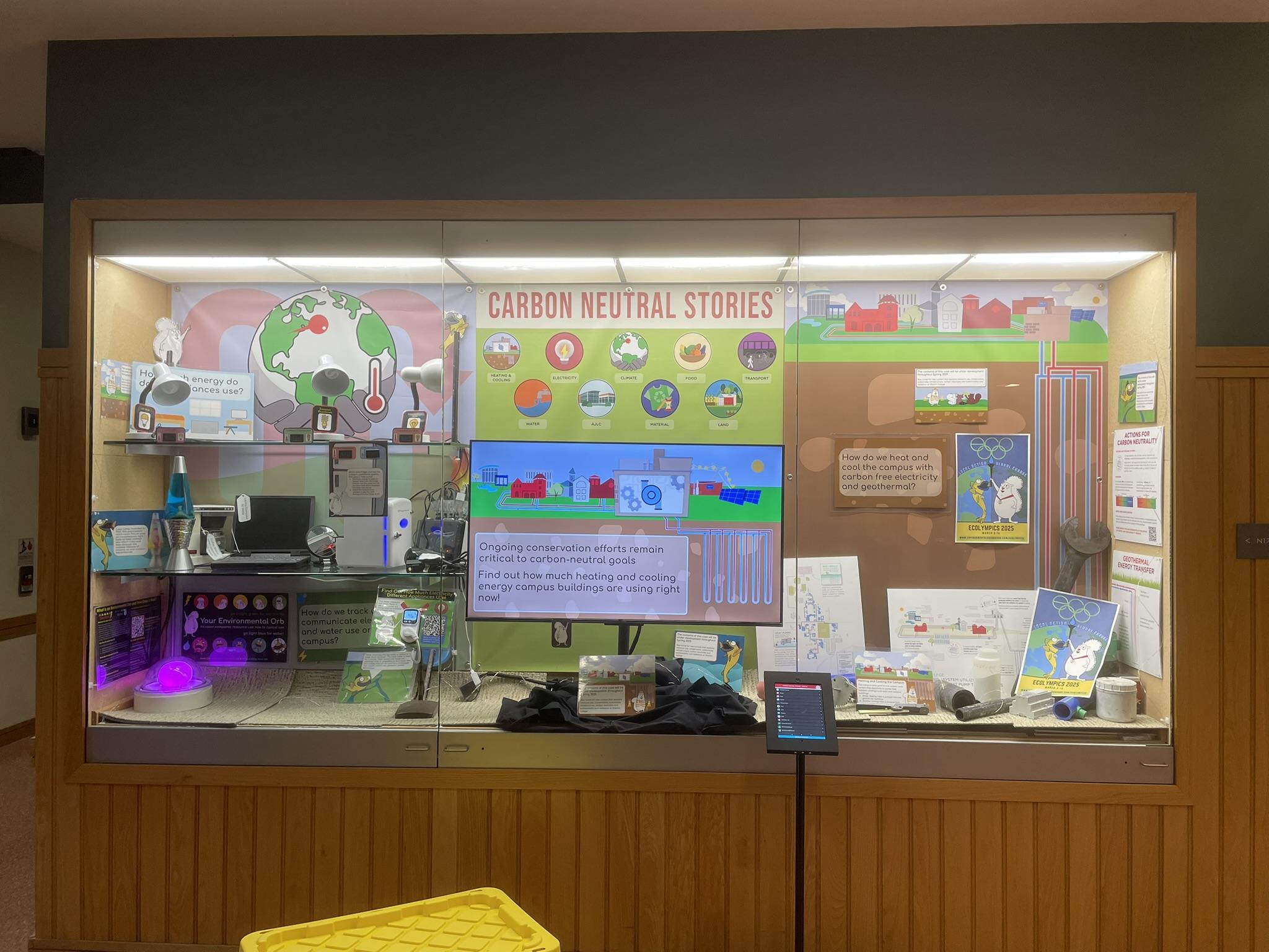 A display case titled Carbon Neutral Stories, with a screen in the middle, posters and objects