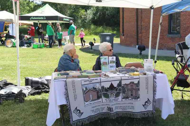 Two people seated at a table with a printed cloth and brochures, under a tent on a lawn